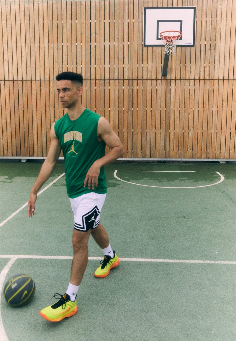 Giocatore di basket con una maglietta senza maniche verde e pantaloni bianchi con dettagli neri, indossando scarpe gialle brillanti mentre gestisce un pallone da basket su un campo.