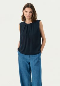 Navyblaue ärmellose Bluse mit gefalteten Details am Ausschnitt, kombiniert mit hellblauen, weiten Jeanshosen. Glatte Stoffoberfläche.