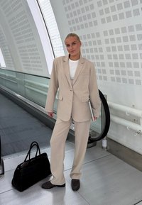 Femme en costume beige se tenant près d'un escalator dans un bâtiment moderne, tenant un ordinateur portable, avec un sac noir posé au sol à côté d'elle.