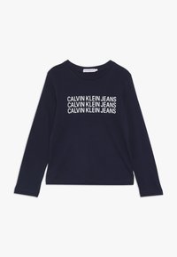 T-paita, jossa pitkä hiha, tummansinistä puuvillaa, jossa "CALVIN KLEIN JEANS" -logo painettuna valkoisena. Klassinen pyöreä pääntie ja rento istuvuus.