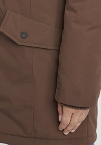 Brun isolert jakke med strukturert stoff, med en fremtredende knappelukking og tettsittende mansjetter. Nærbilde av armen og hånden.