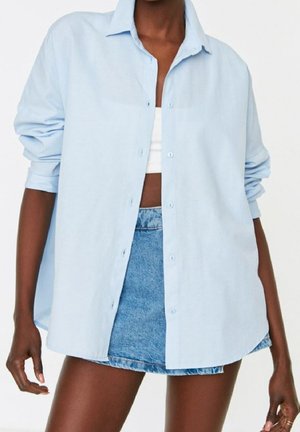 Osoba ubrana w jasnoniebieską, dużą koszulę na guziki założoną na biały top odsłaniający brzuch, w połączeniu z niebieską mini spódniczką dżinsową na jednolitym tle.