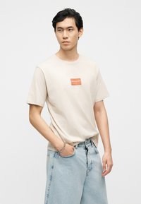 Άνδρας που φοράει μπεζ μπλουζάκι Jack & Jones και ανοιχτόχρωμα τζιν, με το ένα χέρι στην τσέπη και ουδέτερη έκφραση.