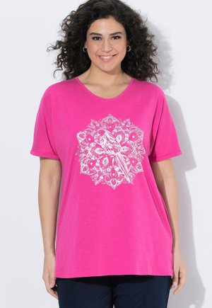 Uśmiechnięta kobieta z kręconymi włosami, ubrana w jasnoróżowy T-shirt z białym ptakiem i wzorem mandali z pawim piórem.