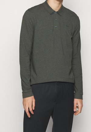 Homme portant un polo à manches longues gris foncé avec deux boutons défaits et un pantalon noir, sur fond uni.