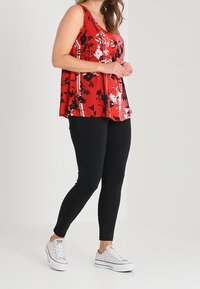 Rotes florales ärmelloses Oberteil mit schwarzen Akzenten, kombiniert mit schwarzen Leggings mit seitlichen Steppdetails und weißen Sneakern.