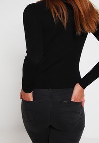 Pitkien punaruskeiden hiusten nainen pukeutuneena mustaan ribbineulepuseroon ja tummiin farkkuihin, kädet takataskuissa, kasvot poispäin kamerasta.