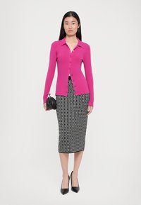 Fuchsfarbener gerippter Cardigan mit Knopfverschluss, kombiniert mit einem schwarz-weißen gemusterten Bleistiftrock. Modell hält eine kleine schwarze Handtasche.