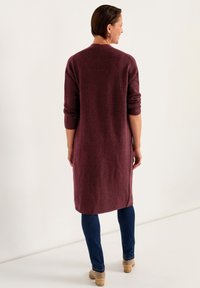Lång vinröd cardigan med strukturstickade detaljer, rund halsringning och uppvikta ärmar, kombinerad med mörkblå jeans och beige klackstövlar.