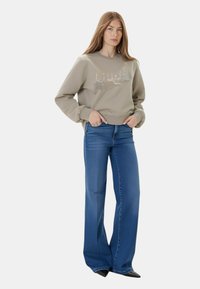 Felpa beige con dettagli grafici argento. Abbinata a jeans a zampa d'elefante blu. L'outfit presenta una vestibilità rilassata e polsini arricciati.