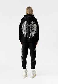 Sudadera negra con diseño de alas metálicas en la parte trasera, combinada con pantalones negros con acentos desgastados y estampados blancos. Botas blancas.