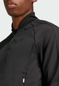 Čierna bomber bunda s hladkým povrchom, so zapínaním na zips, stojatým golierom a detailom loga na hrudi.