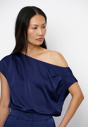 Vrouw met lang zwart haar die een marineblauwe satijnen blouse met één schouder en bijpassende broek met hoge taille draagt, kijkt naar de zijkant.