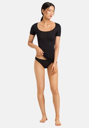 Femme debout pieds nus, portant un haut noir à manches courtes et encolure dégagée ainsi qu’un sous-vêtement style bikini noir, remontant légèrement son t-shirt sur un fond uni.