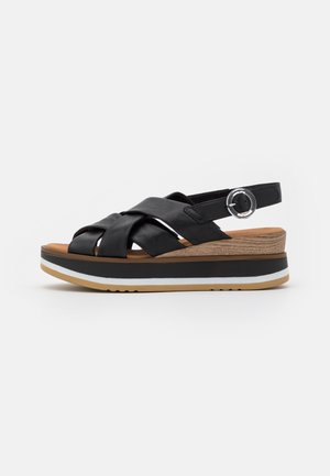 Czarne skórzane sandały z plecioną górą, regulowanym paskiem i srebrnymi akcentami. Posiadają platformę z teksturowanym wyglądem drewna i białymi dodatkami.