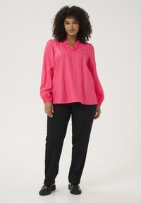Chemisier rose vif avec un col froncé et de longues manches bouffantes ; associé à un pantalon noir à jambe droite et des chaussures noires.