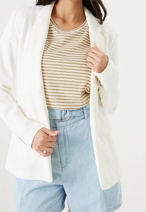 Femme portant un blazer blanc sur un haut rayé beige et blanc avec un short bleu clair taille haute et une ceinture en tissu.
