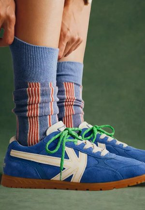 Persoon trekt blauwe geribbelde sokken met rode en witte strepen op, draagt blauwe sneakers met groene veters en bruine zolen op een groene vloer.