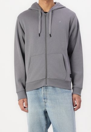 Muž ve šedé mikině na zip s předními kapsami a šňůrkami, v kombinaci se světle modrými džínami, na jednobarevném pozadí.