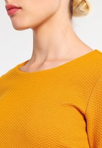 Mosterdgele geribbelde gebreide top met een bootnek en textuurstof. Close-up weergave toont het schouder- en nekgebied en benadrukt het ontwerp.