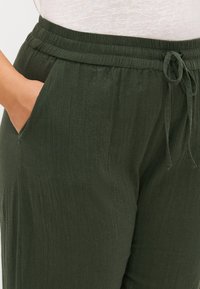 Pantalones ligeros de color verde oscuro con cintura elástica, cordón ajustable y bolsillos laterales; tejido texturizado con líneas verticales sutiles.