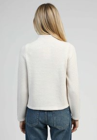 Pull blanc texturé avec un col montant et des manches longues, doté d'un motif gaufré, associé à un jean en denim bleu.