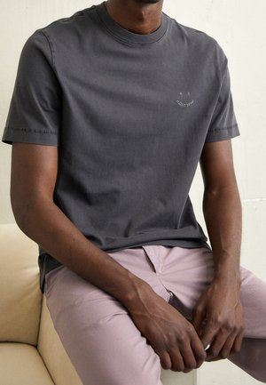 Homme portant un t-shirt gris foncé à manches courtes avec un petit texte blanc sur la poitrine et un pantalon rose clair, assis sur un canapé beige.