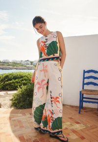 Jeune femme souriante, portant un haut sans manches à motifs floraux et marins ainsi qu'un pantalon large, debout à l'extérieur près d'une chaise bleue au bord de la mer.