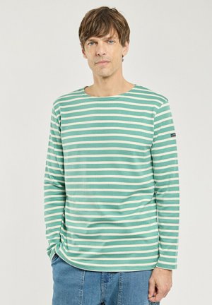 Homme portant une chemise à manches longues rayée verte et blanche et un jean bleu, debout avec une main dans la poche devant un fond uni.