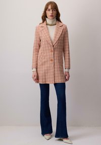 Károvaný kabát v oranžovo-bielej látke, s upraveným strihom, dvomi gombíkmi, klopovým golierom a bočnými vreckami, skombinovaný s modrými rozšírenými džínsami.