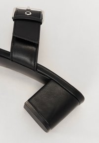 Svart läderklackad sandal med en kraftig klack, med en bred rem och en metallisk spänneaccent. Slät textur med matt yta.