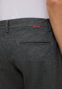Grau strukturierte Hosen mit geradem Schnitt, ausgestattet mit einer Gesäßtasche und einem kleinen roten Etikett. Der Stoff hat ein dezentes Muster und eine glatte Oberfläche.