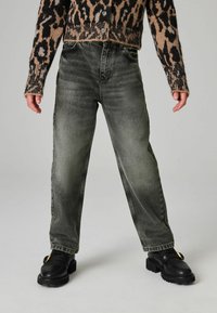 Vaqueros verdes desgastados con corte recto, cinco bolsillos y sutiles desgastes. Combinados con zapatos negros robustos con detalles dorados.