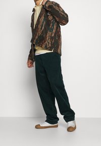 Giacca in corduroy marrone e verde a fantasia su un hoodie giallo chiaro, abbinata a pantaloni in corduroy verde scuro e sneaker bianche con suole marroni.