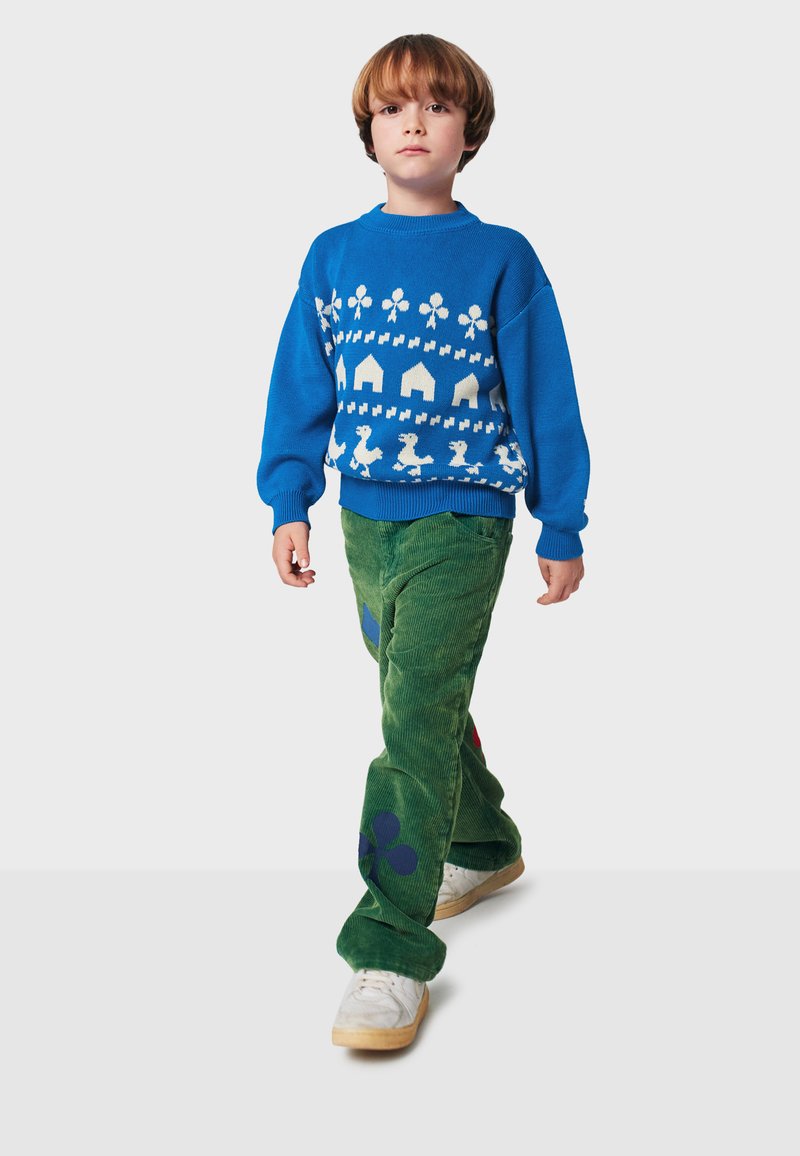 Modrá pletená mikina s bielymi vzormi, zelené kordové nohavice s farebnými tvarmi a biele tenisky. Modelka kráčajúca pred neutrálnym pozadím.