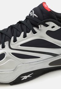 Chaussure de sport noire avec un dessus en mesh respirant, des accents argentés, une semelle en caoutchouc texturé et un système de laçage détaillé.