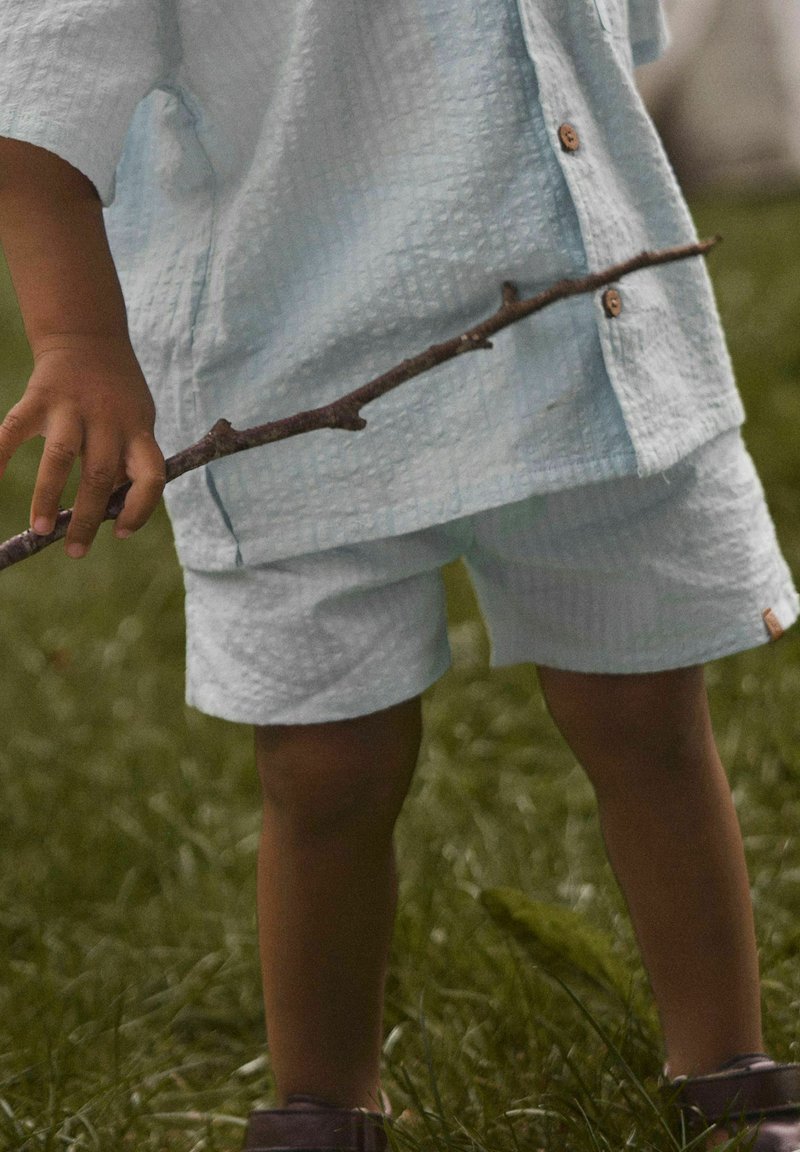 Enfant portant une tenue texturée bleu clair, tenant un bâton, debout sur l'herbe à l'extérieur.