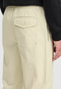 Beige, lichte cargobroek met een knoop achterzak, elastische tailleband en zijnaad. Vloeibare textuur, minimalistisch ontwerp.