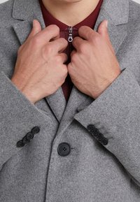 Abrigo de lana gris con botones negros y una capa interior burdeos con cremallera visible. Primer plano de manos ajustando el abrigo en el cuello.