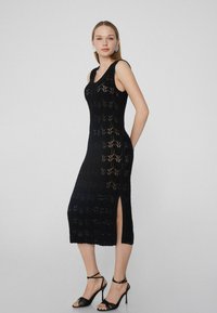 Schwarzes gestricktes Kleid mit einem taillierten Oberteil, seitlichem Schlitz und Zickzackmuster. Kombiniert mit schwarzen Riemchen-High-Heels.