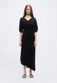 Γυναίκα που στέκεται φορώντας μαύρο μίντι φόρεμα με μανίκια μέχρι τον αγκώνα και μαύρα σανδάλια ανοιχτής μύτης, πάνω σε ένα απλό λευκό φόντο.