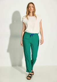 Gröna mönstrade byxor med elastisk midja och blå dragsnor, tillsammans med en enkel vit kortärmad topp och svarta sandaler.