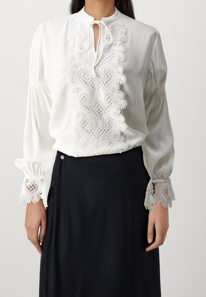 Kobieta ubrana w białą bluzkę z misternymi koronkowymi zdobieniami z przodu i na mankietach, w połączeniu z czarną spódnicą.