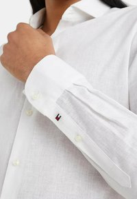 Camicia bianca di lino con colletto classico, caratterizzata da maniche lunghe e polsini con bottoni. Logo rosso, bianco e blu ricamato sul polsino.