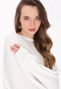 Witte oversized trui met geribde mouwen, zachte textuur en losse pasvorm. Model heeft golvend haar en draagt een ronde oorbel.