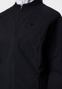 Svart jacka i lättviktsmaterial med frontdragkedja, hög krage och diskret logotypdetalj på bröstet. Slät textur.