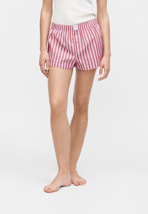 Nő piros-fehér függőleges csíkos pamut boxer rövidnadrágban és fehér ujjatlan felsőben, mezítláb áll fehér háttér előtt.