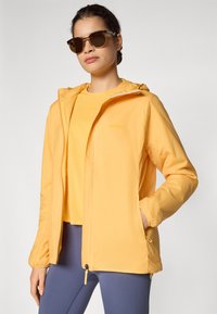 Veste légère jaune avec fermeture éclair sur le devant, poignets élastiques et capuche, portée sur un haut assorti, accompagnée de lunettes de soleil.