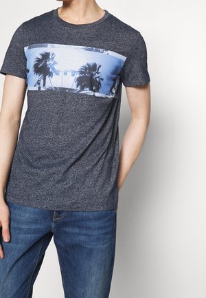 Mand iført mørkegrå T-shirt med blå grafik af palmer og pariserhjul, kombineret med blå jeans, mod hvid baggrund.