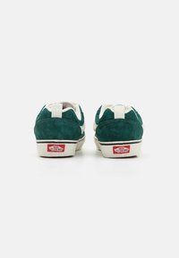 Zapatillas de ante verde con cordones y detalles blancos, que presentan un logo en el talón rojo y un diseño de punta redonda. Suela de goma visible.